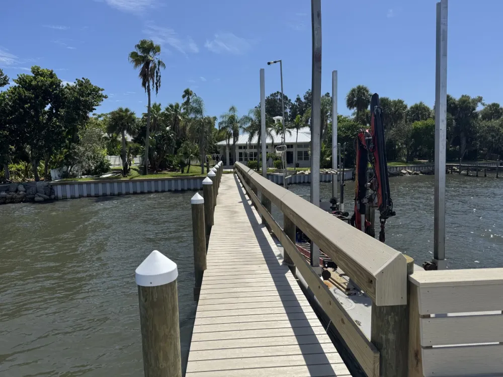 Wooden dock walkway with railings extending over water toward a waterfront home with palm trees